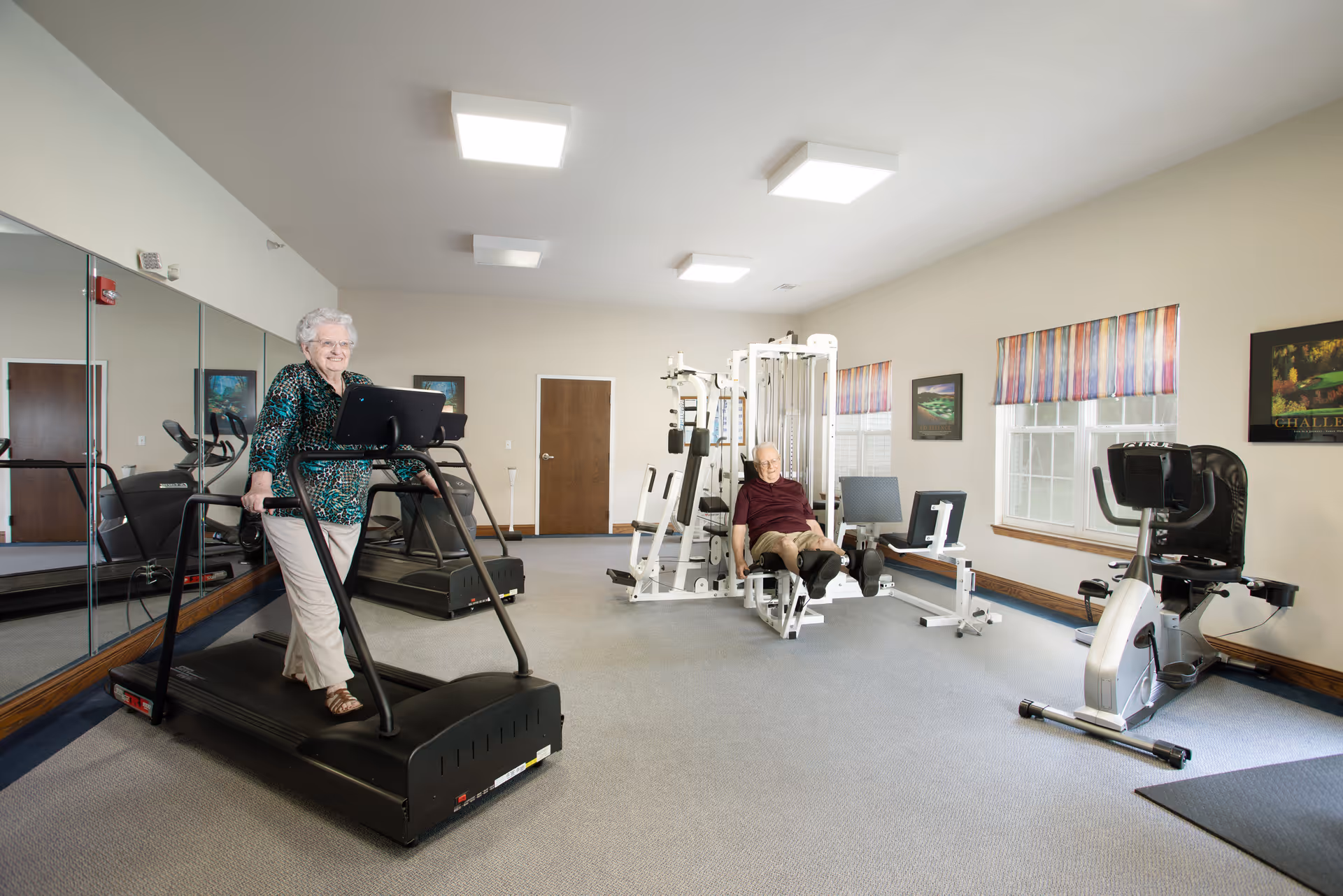 Two elderly residents exercising in a bright fitness room, one woman on a treadmill and a man using a leg machine.