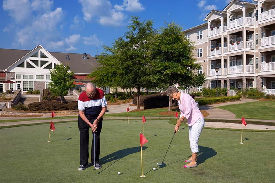 Two elderly people playing golf on a putting green outside a senior living facility with a large building and trees in the background under a blue sky with some clouds.