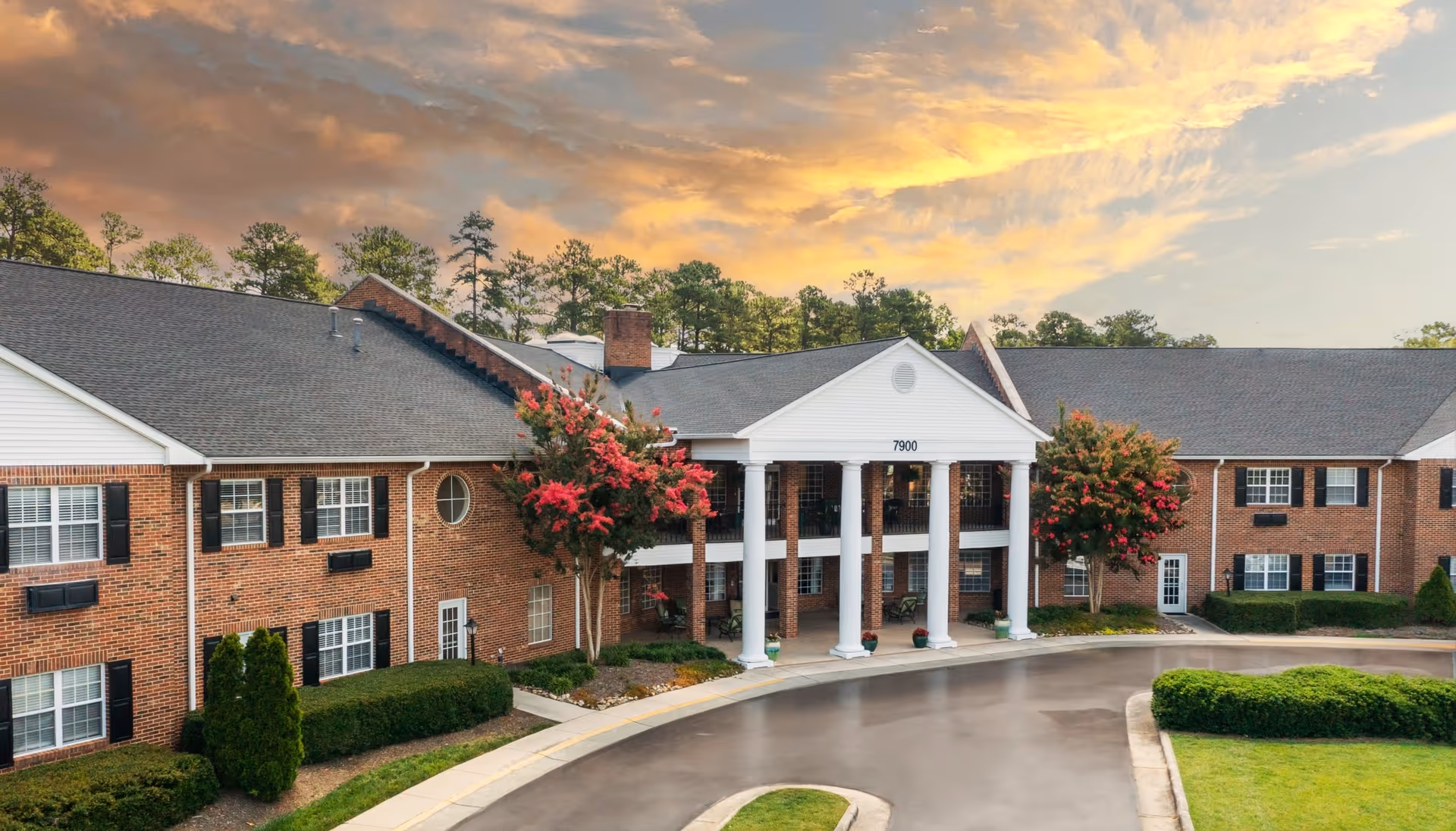 Exterior view of a two-story brick senior living facility with white columns at the entrance, surrounded by well-maintained landscaping including bushes and flowering trees, under a partly cloudy sky at sunset.