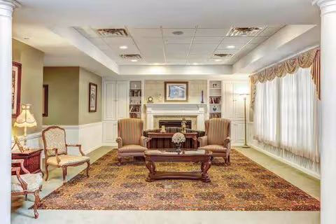 A cozy living room with two striped armchairs and a wooden coffee table on a patterned rug. The room features a white fireplace with built-in shelves on either side, decorated with books and ornaments. Large windows with sheer curtains and valances allow natural light to fill the space. Additional seating includes two upholstered chairs with wooden frames and a side table with a lamp.