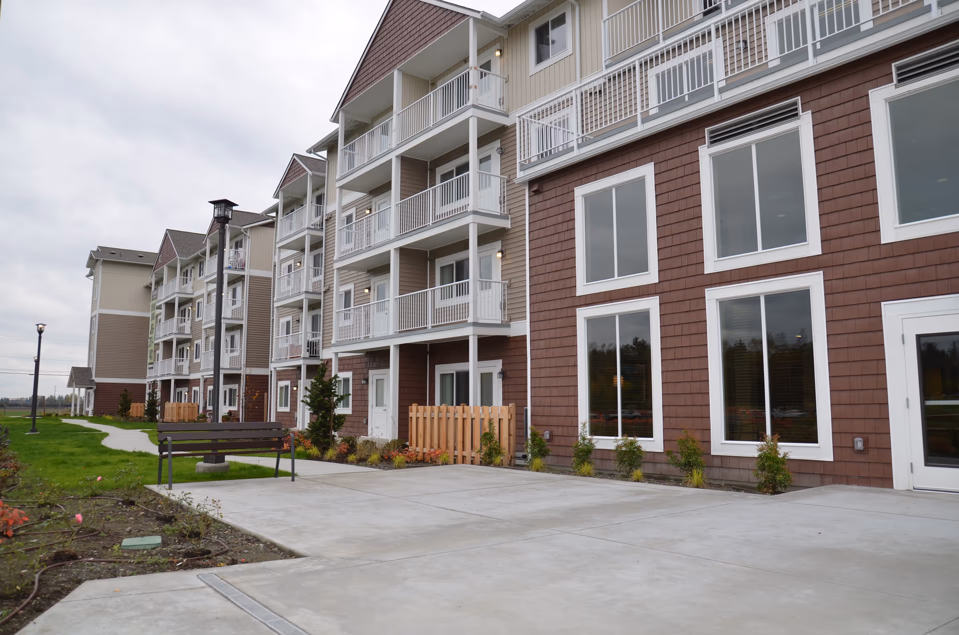 Exterior front of a multi-story senior living building with balconies, large windows, a bench and a landscaped walkway.