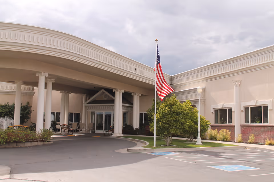 Entrance of a senior living facility with a covered driveway supported by white columns, an American flag on a flagpole, some greenery, and a few benches near the entrance under a cloudy sky.