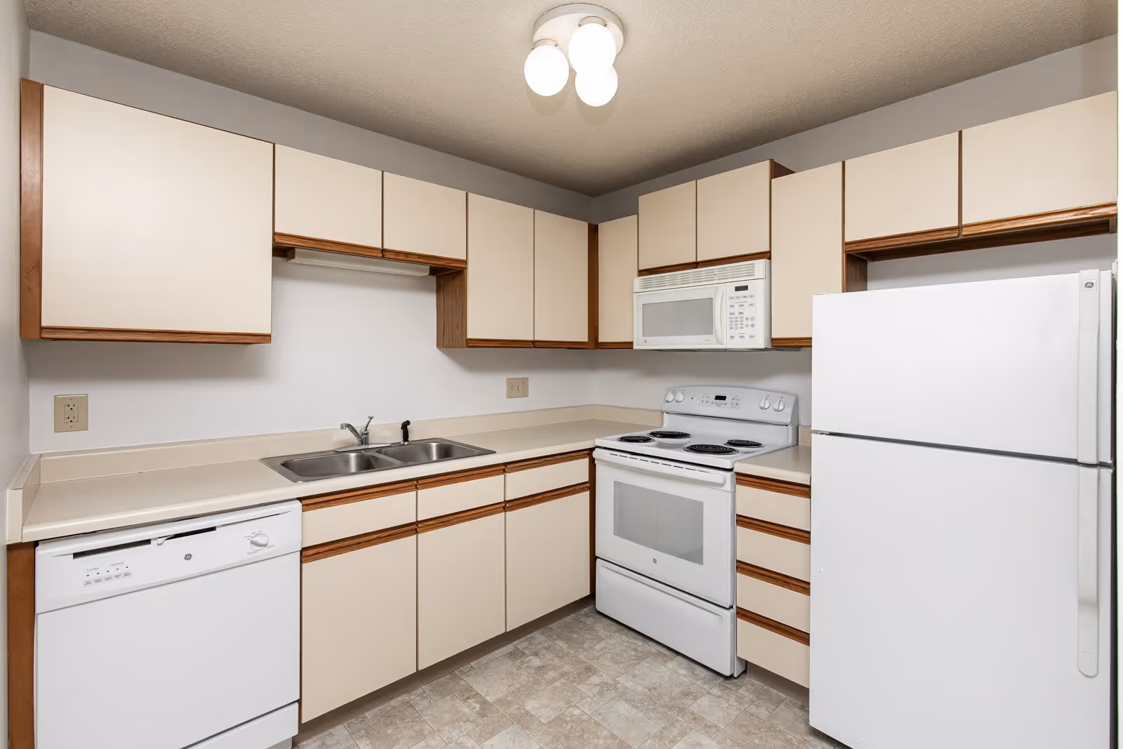 A clean kitchen with beige cabinets and wooden trim, featuring a white dishwasher, double sink, electric stove with oven, microwave, and a refrigerator. The floor has a light-colored tile pattern and the ceiling has a light fixture with three round bulbs.