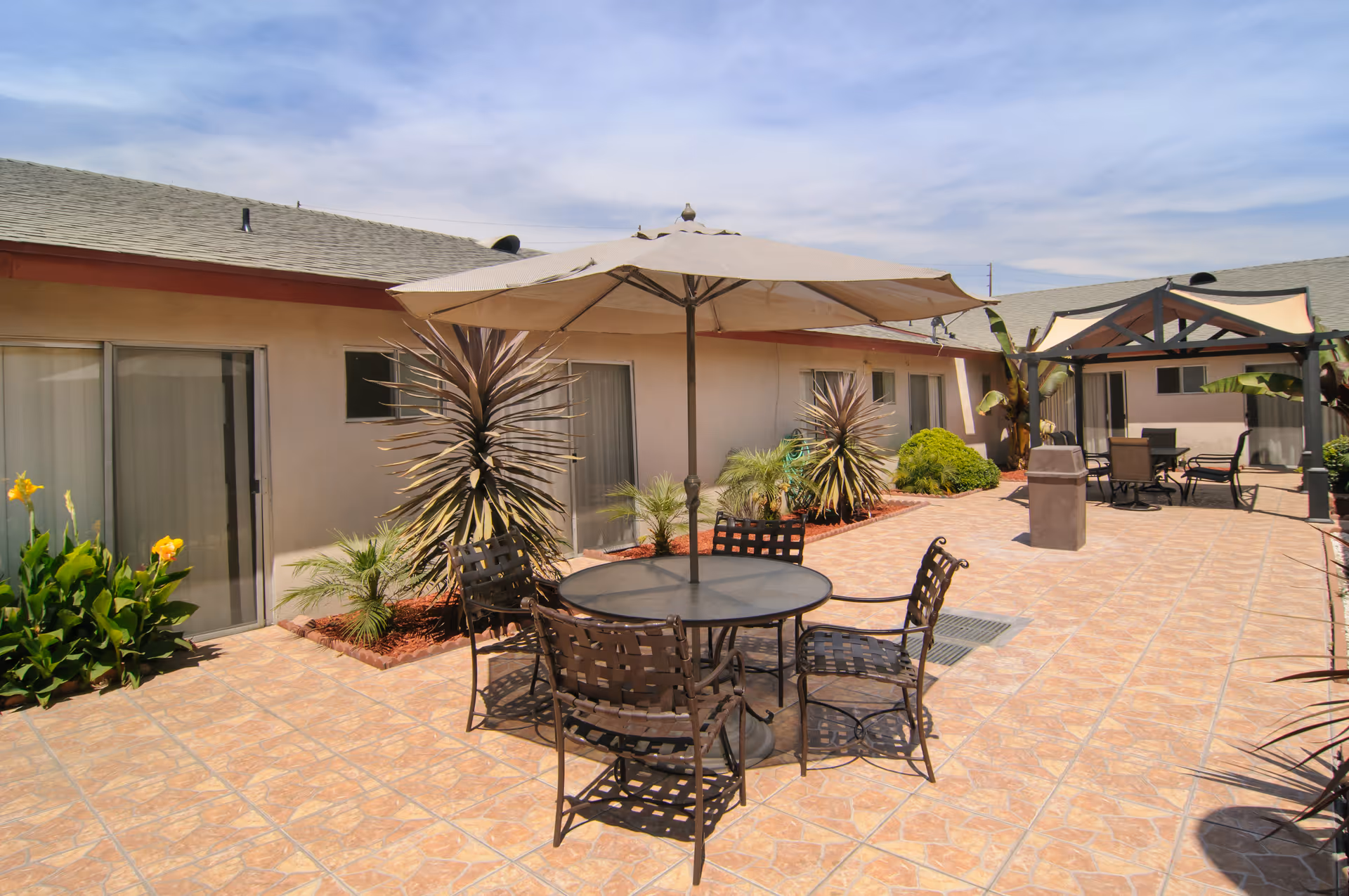 Outdoor patio area at Bel Tooren Villa Convalescent Hospital with tiled flooring, metal table and chairs under a large beige umbrella, surrounded by plants and shrubs, and additional seating under a gazebo structure.