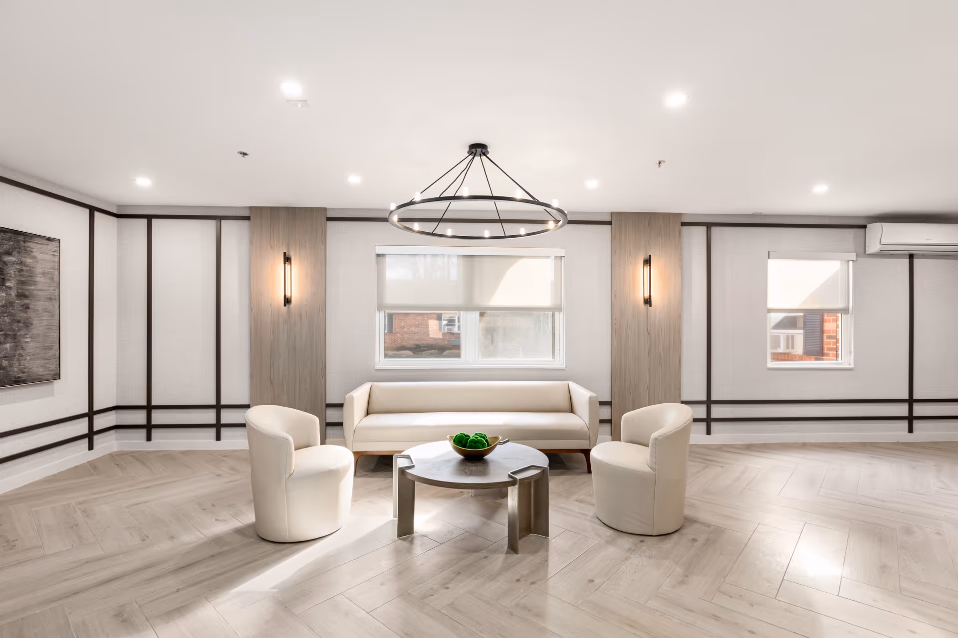 Bright modern lounge with a sofa and three upholstered chairs arranged around a round coffee table beneath a circular chandelier.