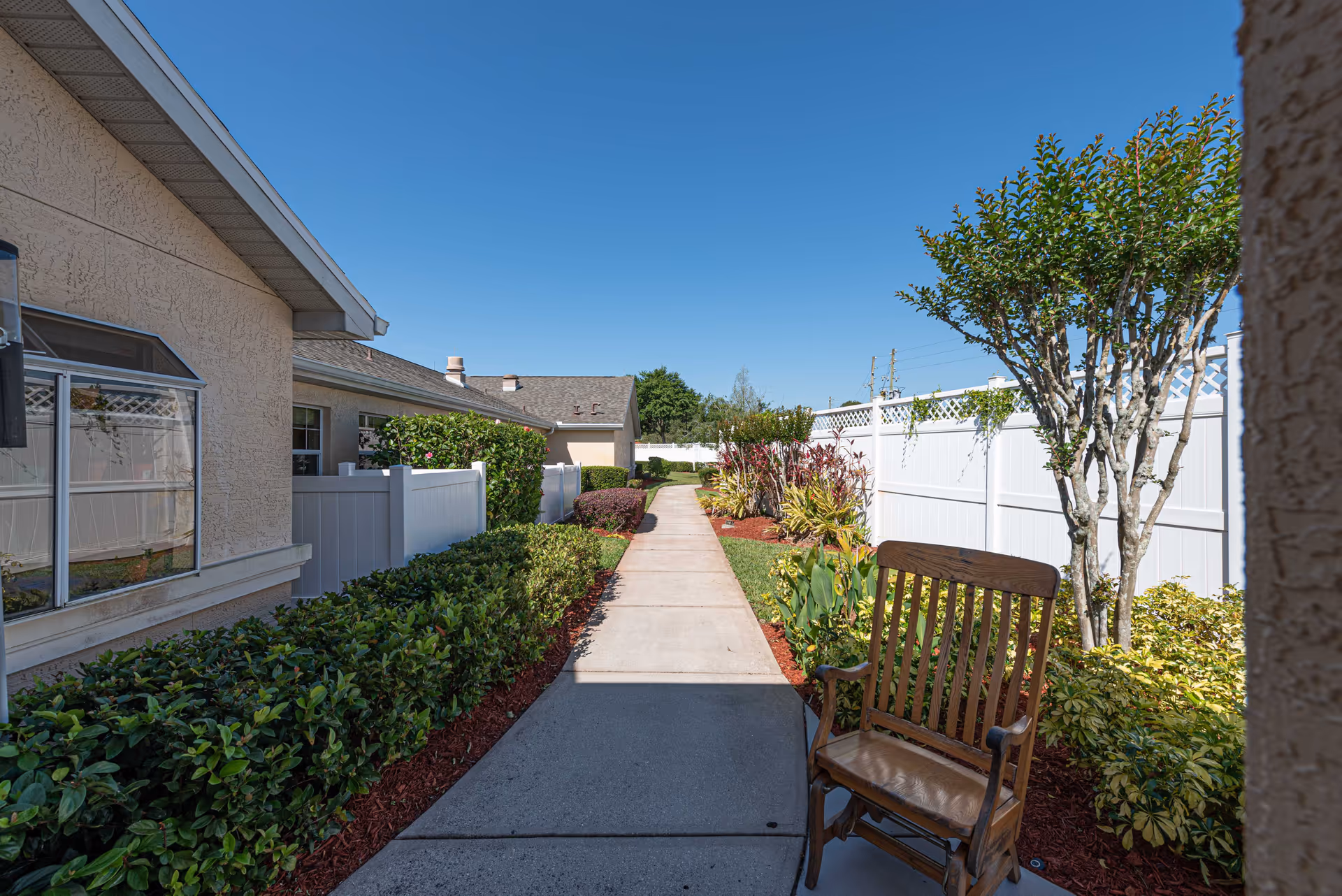 A sunny outdoor walkway at Arden Courts - ProMedica Memory Care Community (Winter Springs) bordered by green bushes, plants, and a white fence. A wooden rocking chair is placed on the right side near a tree.