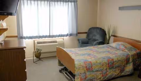 Assisted living resident room with bed, quilt, chair, dresser, window blinds, and plant.