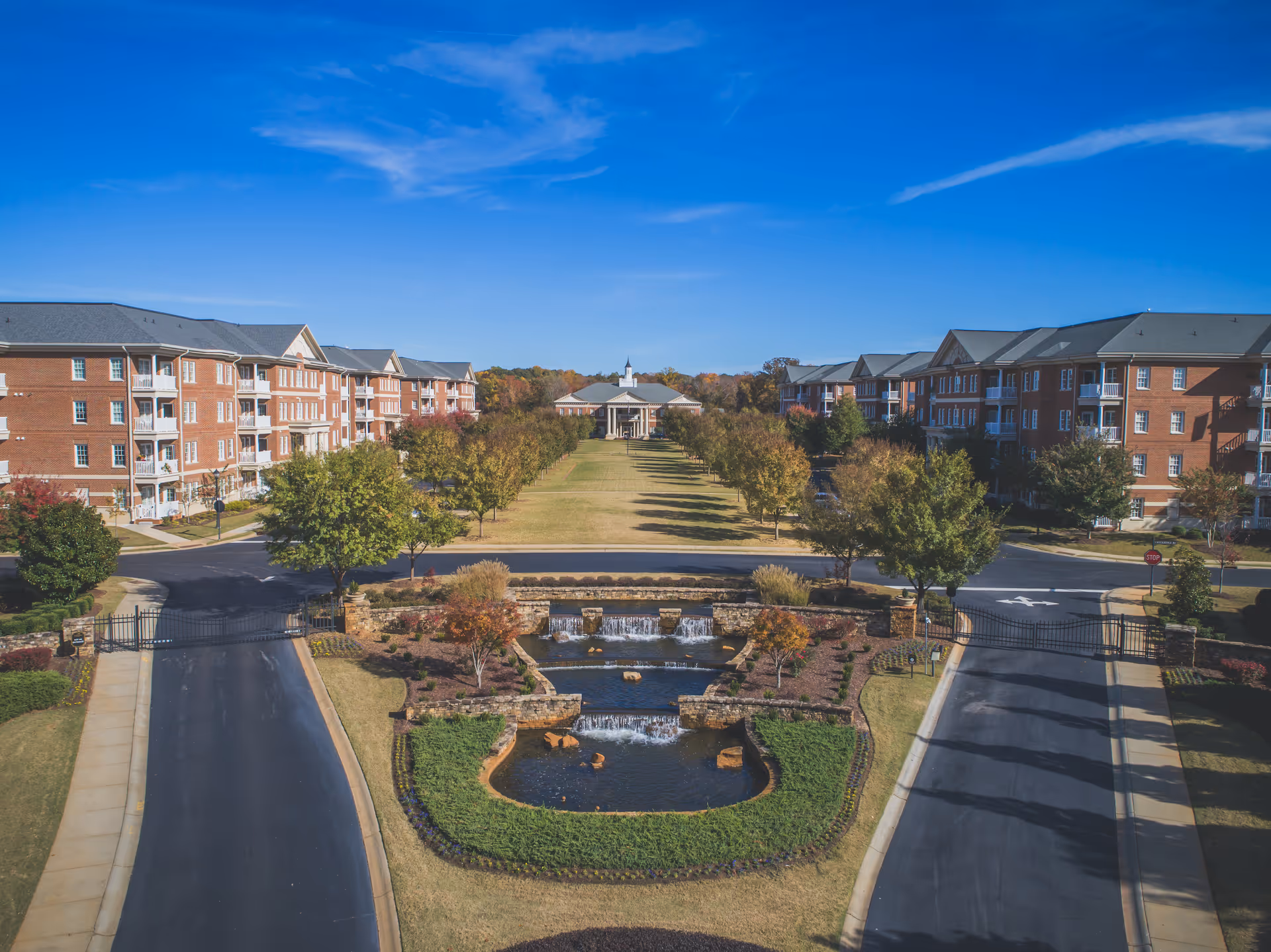 Aerial view of a senior living facility with two large brick buildings on either side of a central landscaped area featuring a tiered water fountain and a long grassy pathway lined with trees leading to a pavilion in the distance under a clear blue sky.
