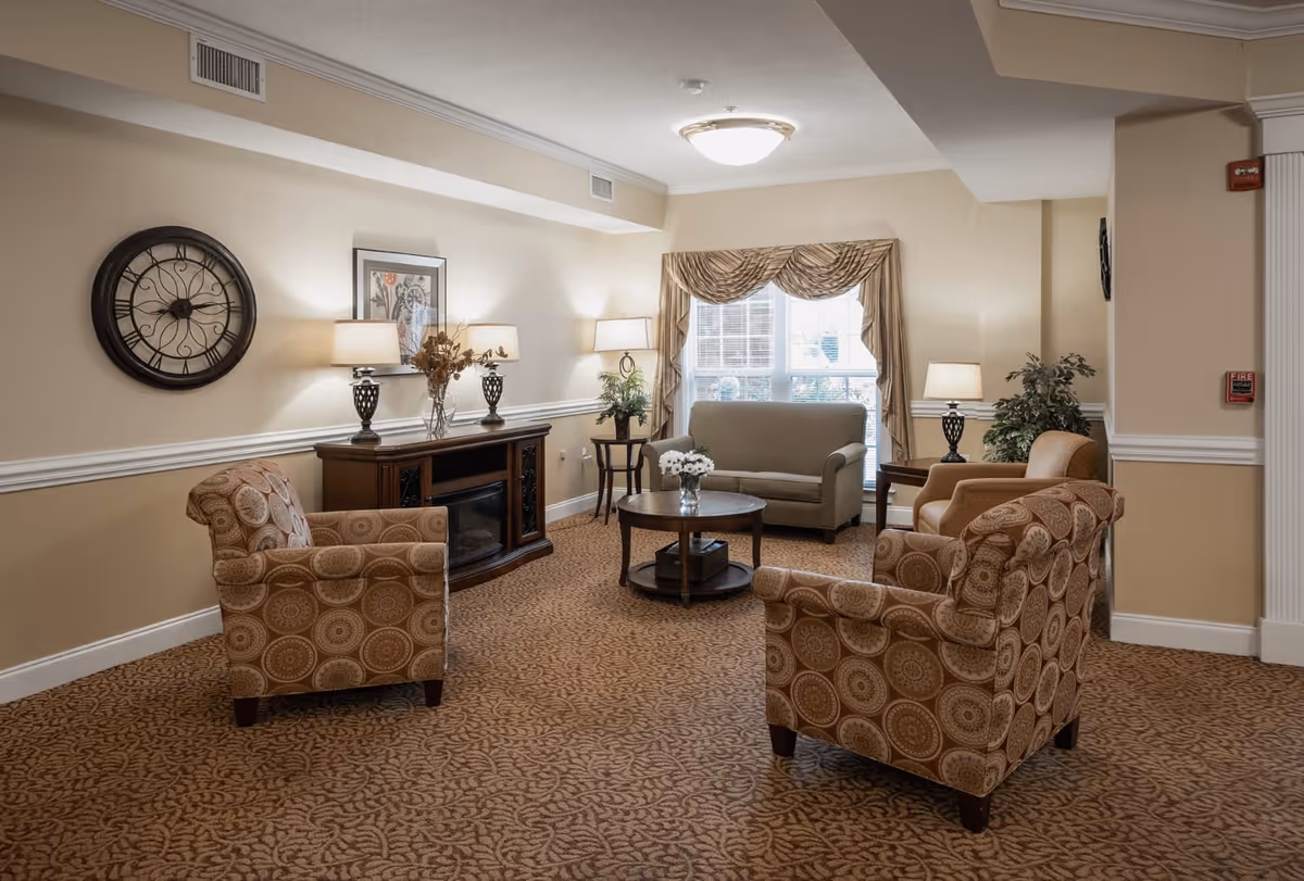 A cozy living room area with beige walls and patterned carpet. The room features a round wooden coffee table with a vase of white flowers, a beige loveseat near a window with draped curtains, and four patterned armchairs arranged around the table. There are three table lamps on wooden side tables and a wooden cabinet with decorative items and a wall clock above it.