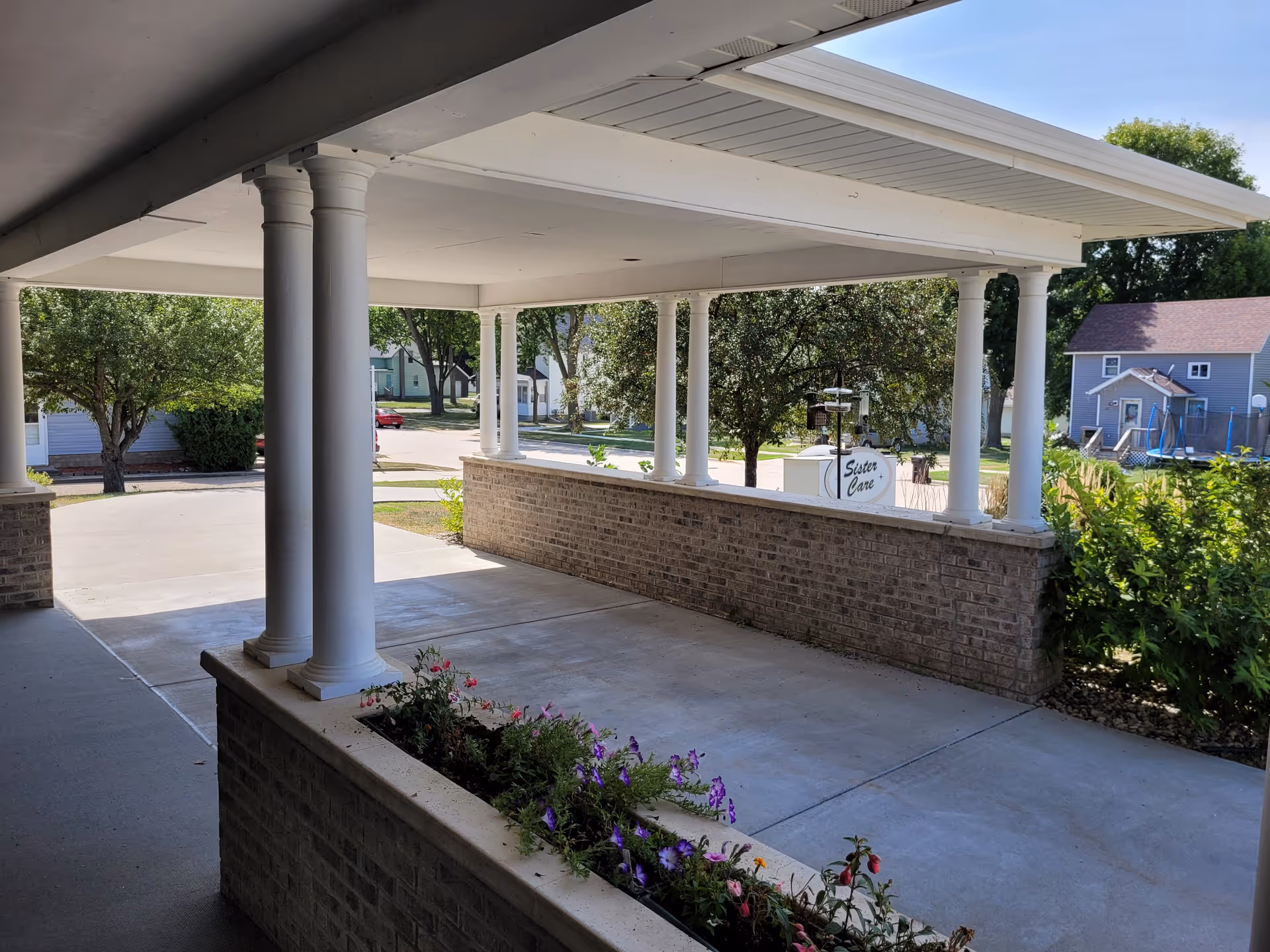 Covered front entrance with white columns, brick half-walls and a flower planter, overlooking a driveway and a sign reading 'Sister Care'.