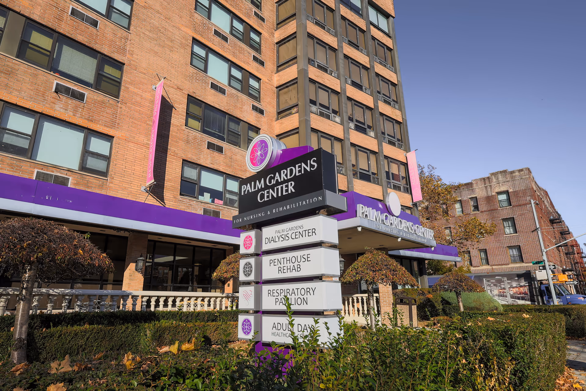 Exterior view of Palm Gardens Center for Nursing & Rehabilitation, a multi-story brick building with large windows and purple signage. There is a signpost in front listing services including Dialysis Center, Penthouse Rehab, Respiratory Pavilion, and Adult Day Healthcare. Shrubs and small trees are visible in the foreground under a clear blue sky.