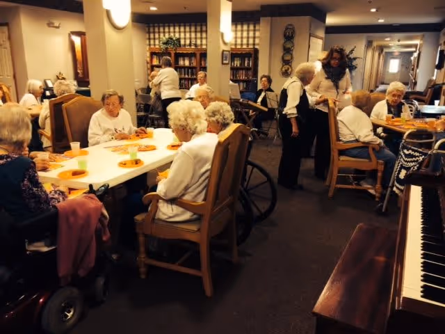 A group of elderly people sitting around tables in a communal room, some in wheelchairs, engaging in conversation and activities. The room has bookshelves, warm lighting, and a piano visible in the foreground.