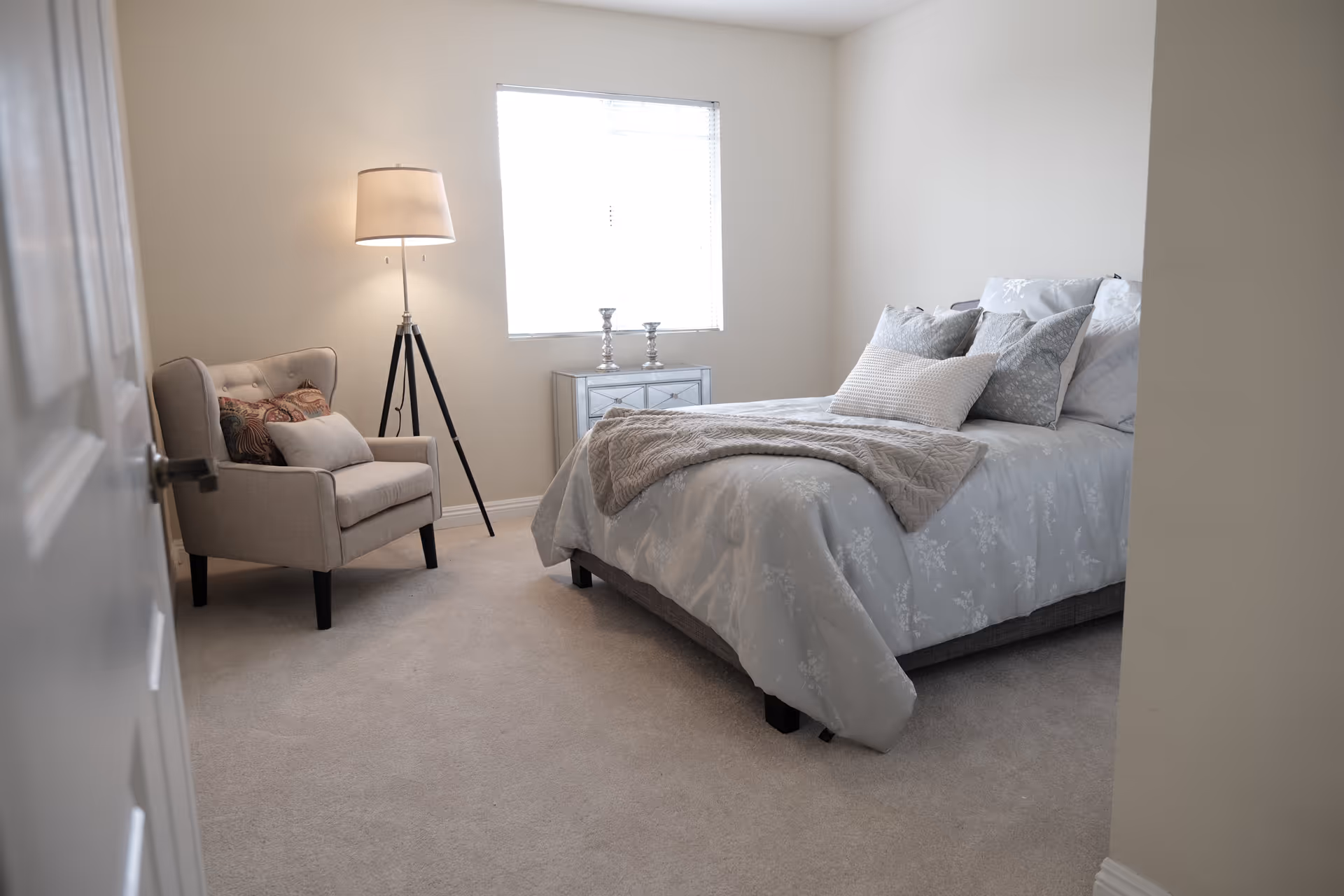 A cozy bedroom with a neatly made bed featuring multiple pillows and a light gray floral comforter. To the left of the bed is a beige armchair with decorative pillows, and a tall floor lamp with a tripod base stands next to the chair. A small mirrored nightstand with two silver candle holders is positioned under a window with blinds, allowing natural light to fill the room. The walls are painted a soft beige color and the floor is carpeted in a light neutral tone.