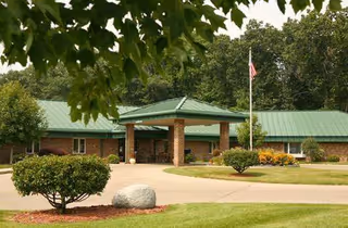 Exterior view of Valley Pines Senior Living facility showing a single-story brick building with a green metal roof, a covered entrance supported by brick columns, a flagpole with an American flag, and landscaped grounds with bushes, trees, and a large rock.