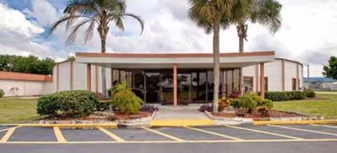 Front of Grand Villa of Lakeland with glass entrance, palm trees, and a parking lot.