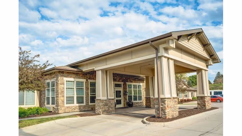Exterior view of Cedar Creek of Prairie Meadows facility entrance with a covered drop-off area supported by stone and beige columns, surrounded by windows and landscaping under a partly cloudy sky.