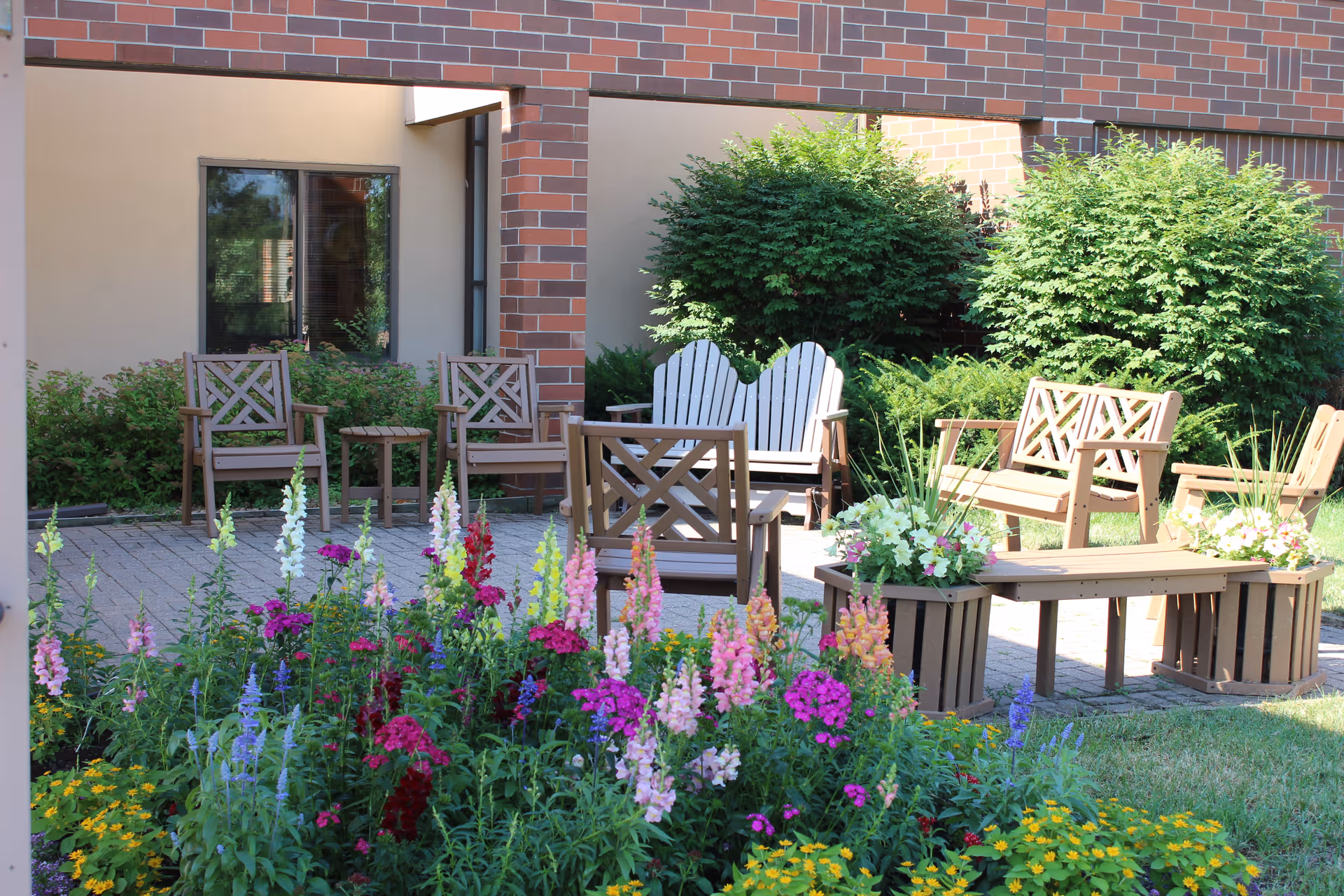 Sunlit courtyard with wooden benches and chairs surrounded by colorful flower beds and shrubs in front of a brick building.