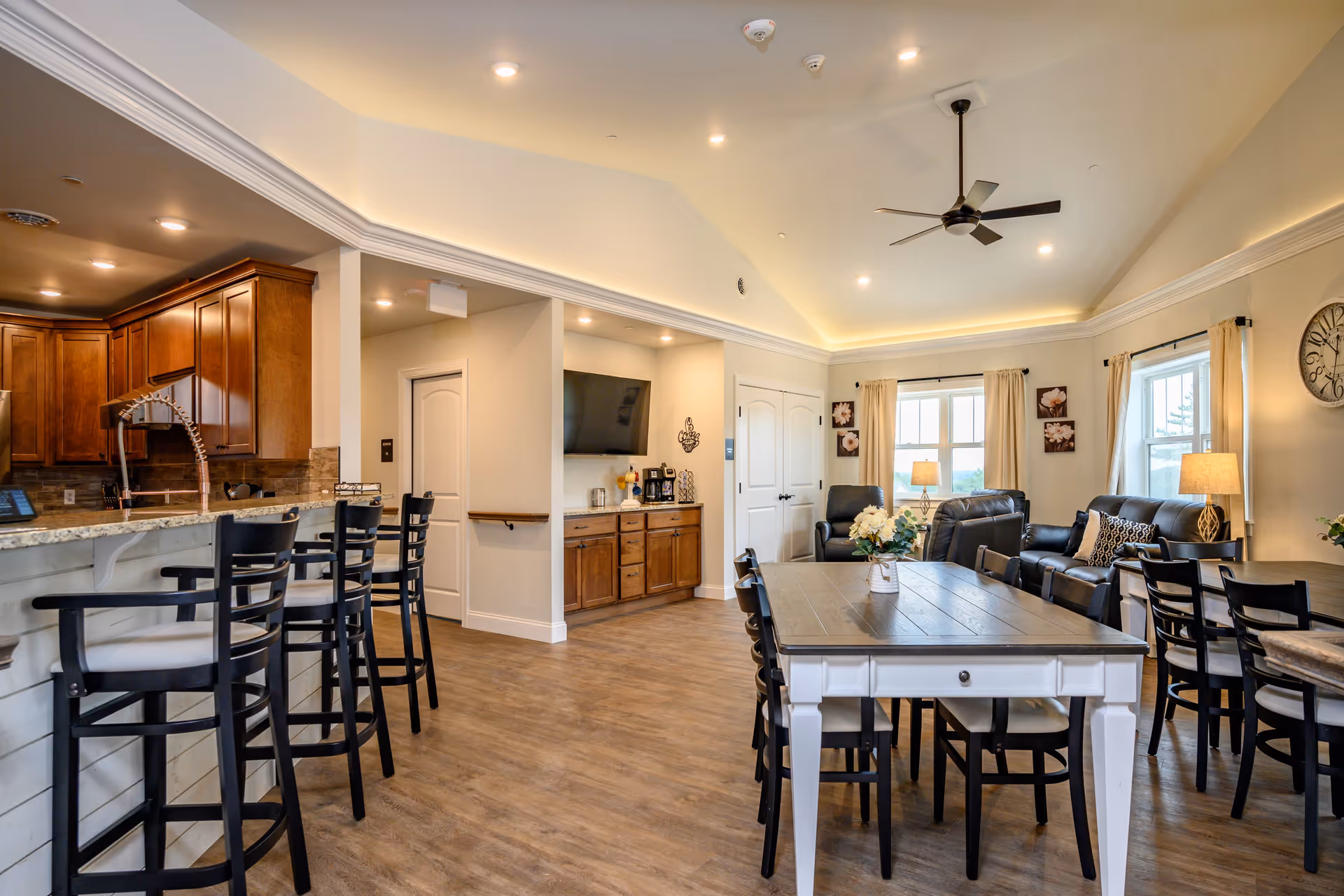 A spacious and well-lit common area in Victor Views Assisted Living featuring a kitchen with wooden cabinets and a granite countertop with bar stools, a dining area with tables and chairs, and a living room area with leather sofas and armchairs. The room has wood flooring, large windows with curtains, ceiling lights, a ceiling fan, and decorative wall art.