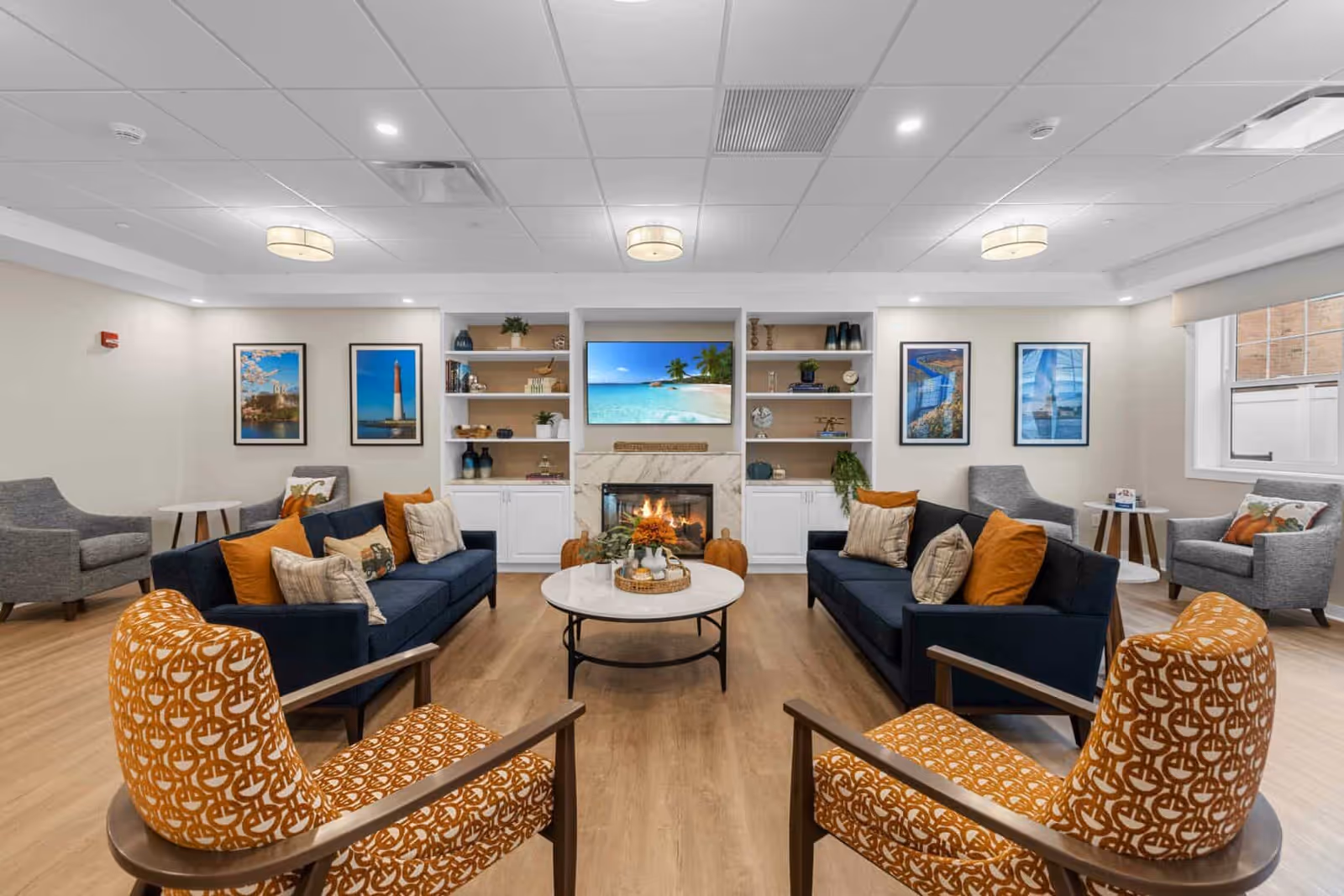 A bright and modern living room area with two navy blue sofas adorned with orange and beige pillows, two patterned orange armchairs, and two gray armchairs near windows. A round white coffee table is centered on a wooden floor. The back wall features a fireplace with a TV above it, flanked by built-in white shelves and cabinets decorated with plants, books, and ornaments. Framed pictures hang on the walls.