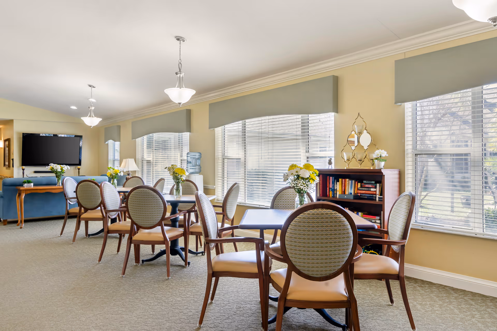 A bright and spacious common area in a senior living facility with multiple round tables and chairs arranged for seating. Each table has a vase with fresh flowers. Large windows with blinds allow natural light to fill the room. A blue couch and a flat-screen TV mounted on the wall are visible in the background. A bookshelf with books and decorative items is placed near the windows.