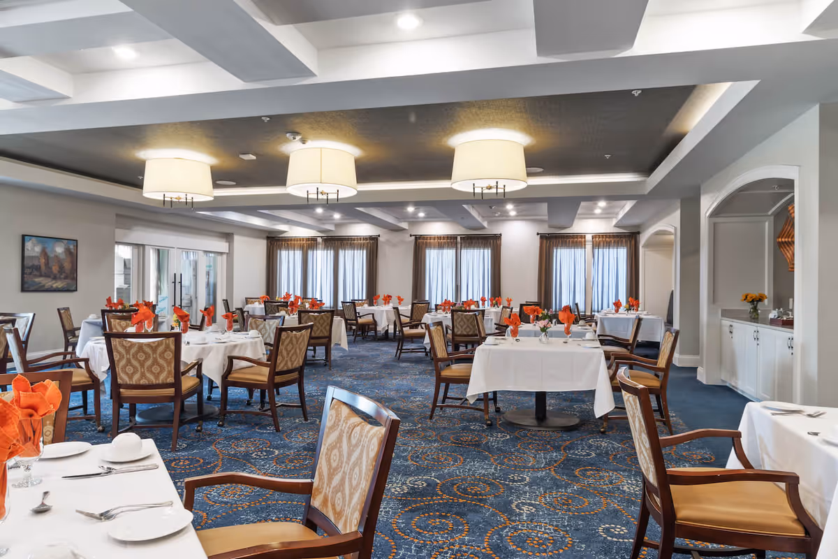 A spacious dining room with multiple round and square tables covered with white tablecloths, each set with plates, silverware, and orange folded napkins. The room features patterned carpet with blue and orange swirls, cushioned chairs with wooden frames, large windows with sheer curtains, and modern ceiling lights.