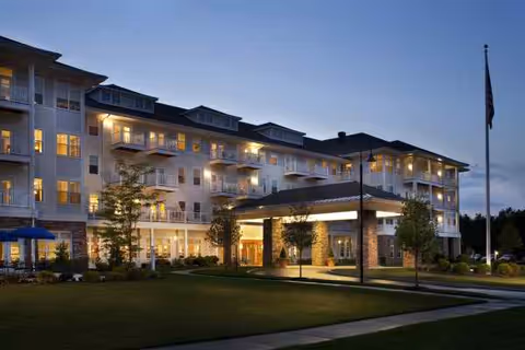 Front exterior of a multi-story senior living building at dusk with illuminated balconies and a covered entrance.