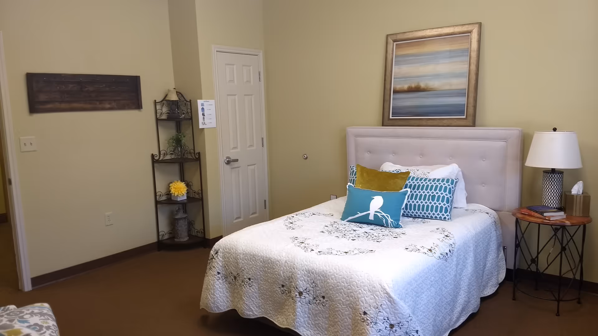 A cozy bedroom with a neatly made bed featuring a white quilt and decorative pillows in blue, green, and white. Above the bed hangs a framed landscape painting. To the right of the bed is a round wooden side table with a lamp, books, and a tissue box. In the corner, there is a metal shelf with decorative items and a closed white door. The walls are painted light beige and the floor is carpeted brown.