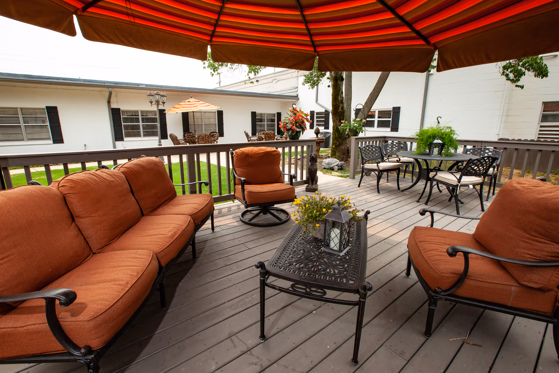 Outdoor patio area with cushioned seating including a sofa and chairs with rust-colored cushions, a black metal coffee table with a lantern and flowers, and a large red umbrella providing shade. In the background, there is a white building with windows and additional seating with tables and chairs under umbrellas.