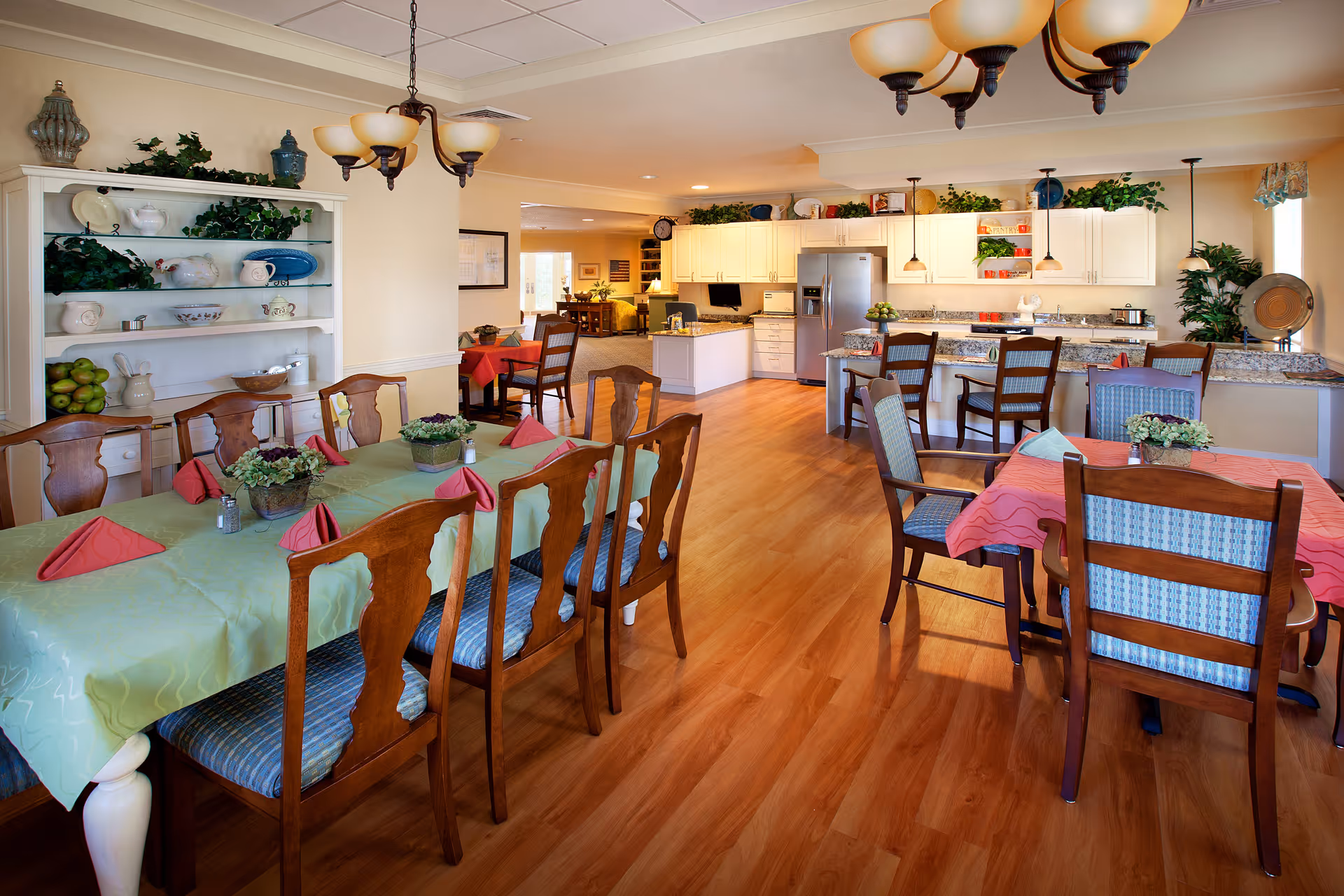 A bright dining area in a senior living facility featuring wooden tables and chairs with colorful tablecloths and folded napkins. The room has a wooden floor, a white cabinet with decorative items and plants, and a kitchen area with white cabinets, granite countertops, and stainless steel appliances in the background.