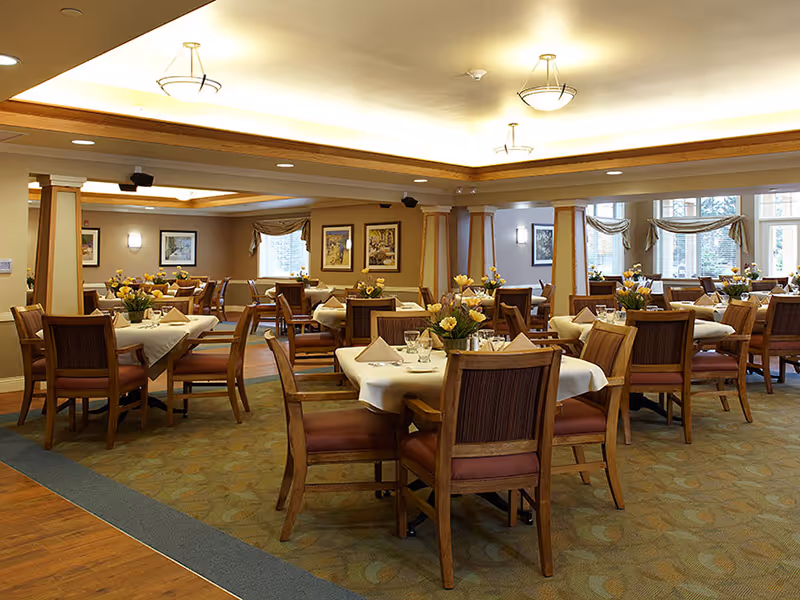 A spacious dining room with multiple tables covered in white tablecloths, each set with napkins, glassware, and floral centerpieces. The room features wooden chairs with padded seats, soft lighting from ceiling fixtures, framed artwork on the walls, and large windows with draped curtains allowing natural light to enter.