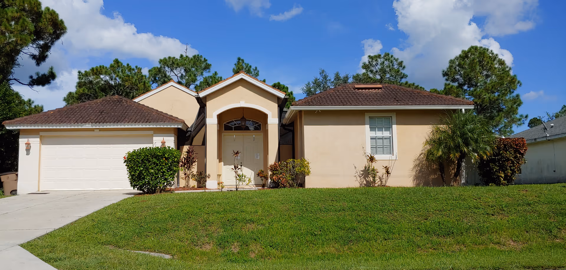Single-story beige house with a brown tiled roof, a white garage door on the left, a central entrance with an arched doorway, and a window on the right side. The house is surrounded by green grass, small bushes, and trees under a partly cloudy blue sky.
