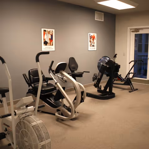 Small exercise room with recumbent bikes, a rowing machine, and framed posters on gray walls.