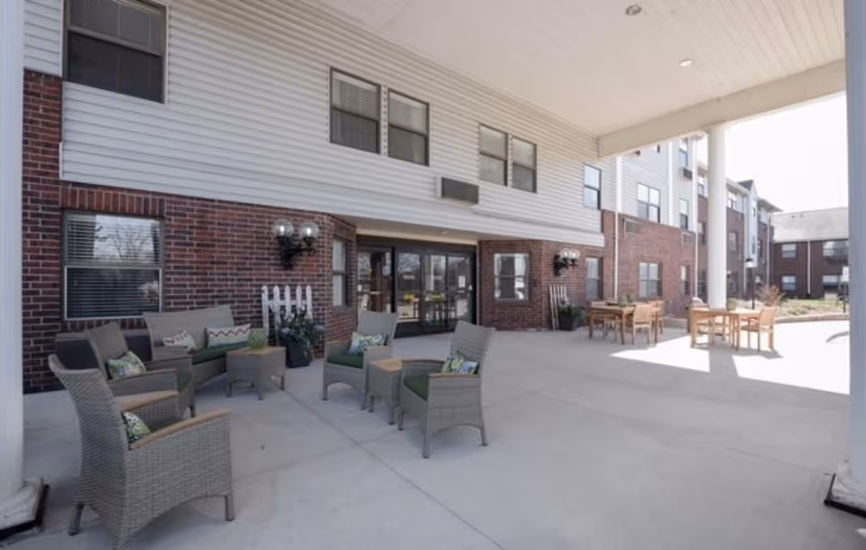 Outdoor covered patio area at Towne Centre with wicker chairs and a small sofa arranged around a coffee table on the left side, and wooden tables with chairs on the right side. The building exterior features red brick and white siding with multiple windows.