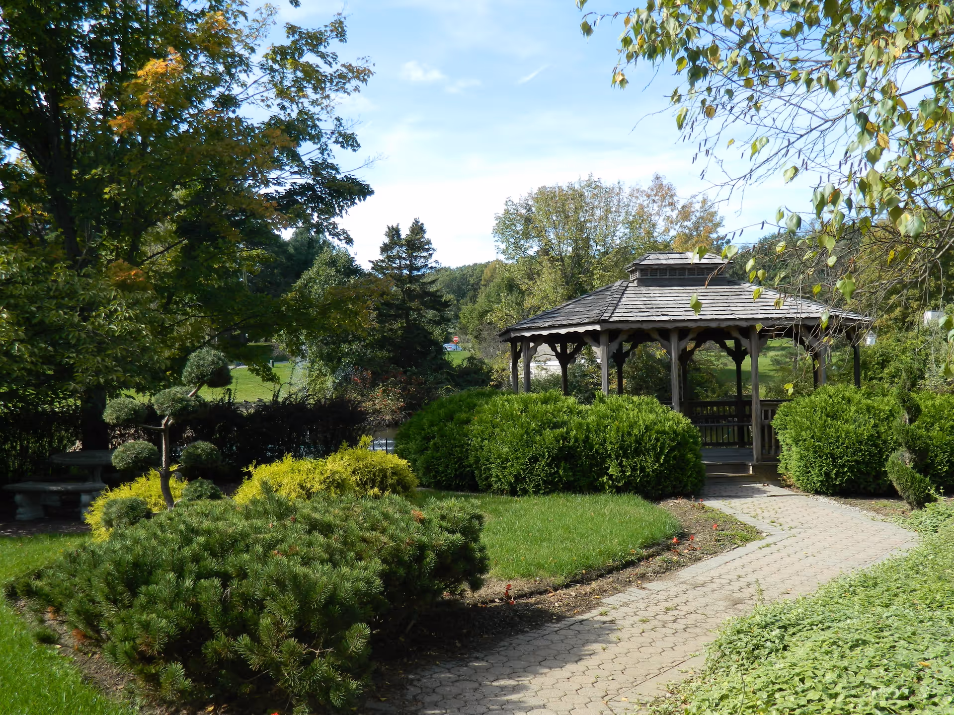 A peaceful outdoor garden area with a paved walkway leading to a wooden gazebo surrounded by green bushes and trees under a clear blue sky.