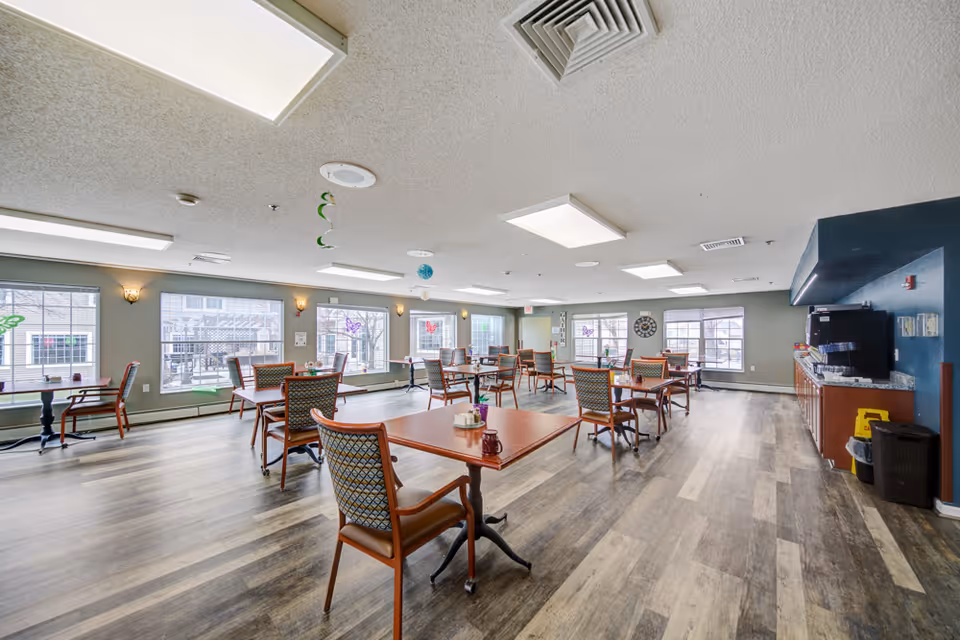 A spacious dining room with multiple wooden tables and cushioned chairs arranged neatly. Large windows allow natural light to fill the room, and there are decorative butterfly decals on the windows. The room has a wood-patterned floor and a ceiling with recessed lighting. On the right side, there is a counter area with a coffee machine and other supplies.