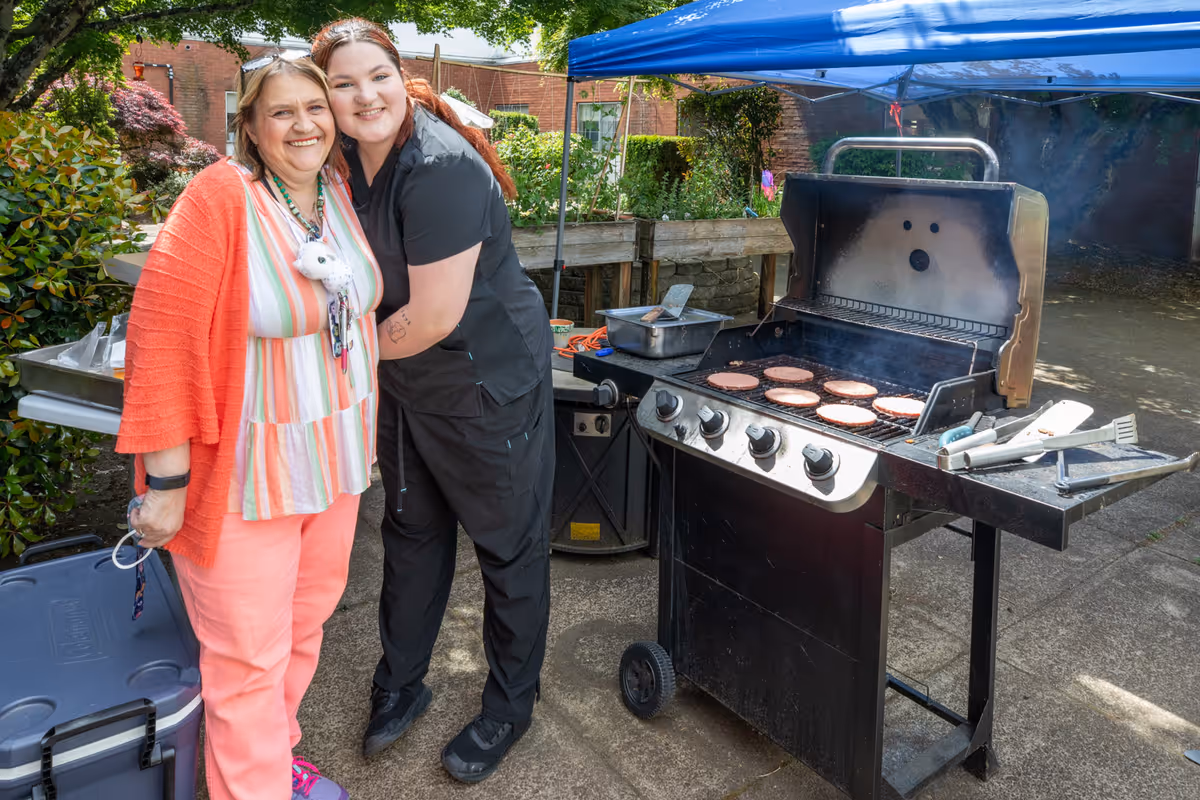 Two women smiling and posing together outdoors next to a barbecue grill with several burger patties cooking. The setting appears to be a garden or patio area with greenery and a blue canopy overhead.