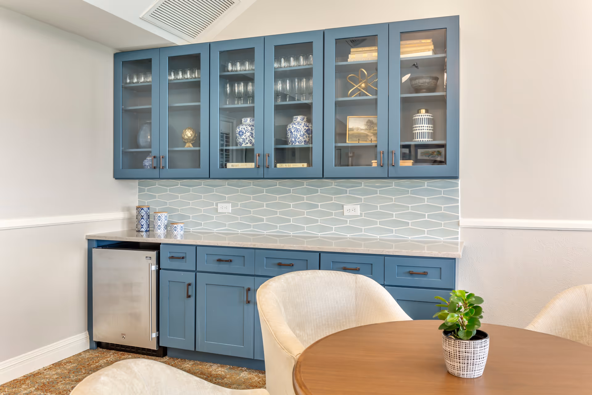 A kitchen area with blue cabinets featuring glass doors displaying glassware and decorative items. Below the cabinets is a light-colored countertop with a light blue hexagonal tile backsplash. There is a small stainless steel refrigerator under the counter. In the foreground, there is a round wooden table with a small potted plant and cream-colored upholstered chairs around it.