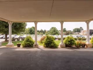 View from a covered entrance or porch area looking out towards a parking lot with several cars and landscaped greenery including bushes and trees. The area is supported by white columns and has a paved driveway.