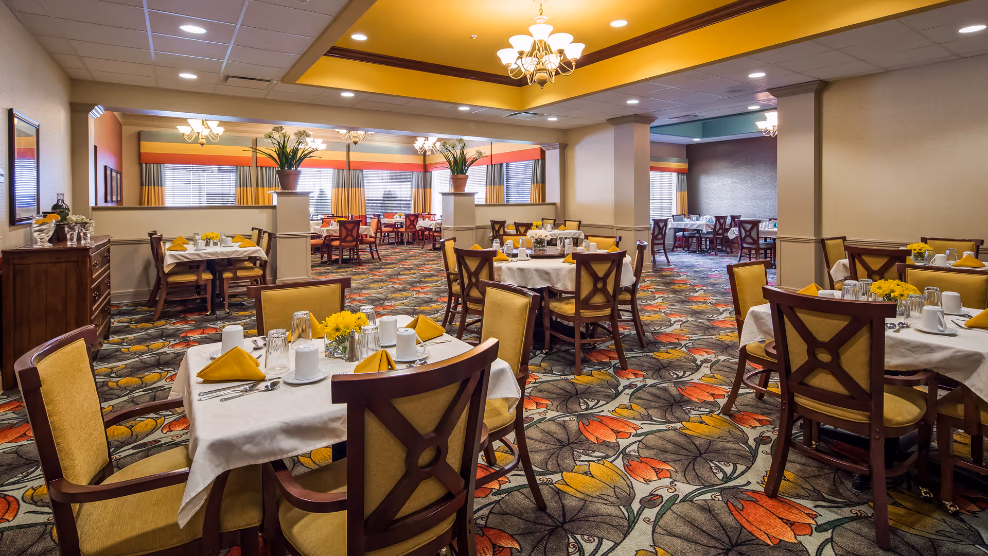 A spacious dining room with multiple tables covered in white tablecloths, each set with yellow napkins, cups, and silverware. The room features patterned carpet with orange, yellow, and gray floral designs, wooden chairs with yellow cushions, and large windows with yellow and orange curtains. There are decorative plants on pillars and chandeliers hanging from the ceiling.