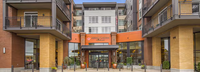 Front entrance of Merrill Gardens at Kirkland, a multi-story senior living facility with brick and beige exterior walls, balconies, large windows, and a covered entryway with potted plants on either side.