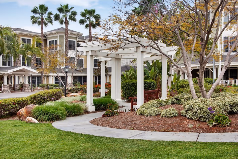 Outdoor garden area at Ivy Park at Seal Beach featuring a white wooden pergola with a bench underneath, surrounded by landscaped greenery, bushes, trees, and a curved concrete pathway. The multi-story residential building with large windows and palm trees is visible in the background.