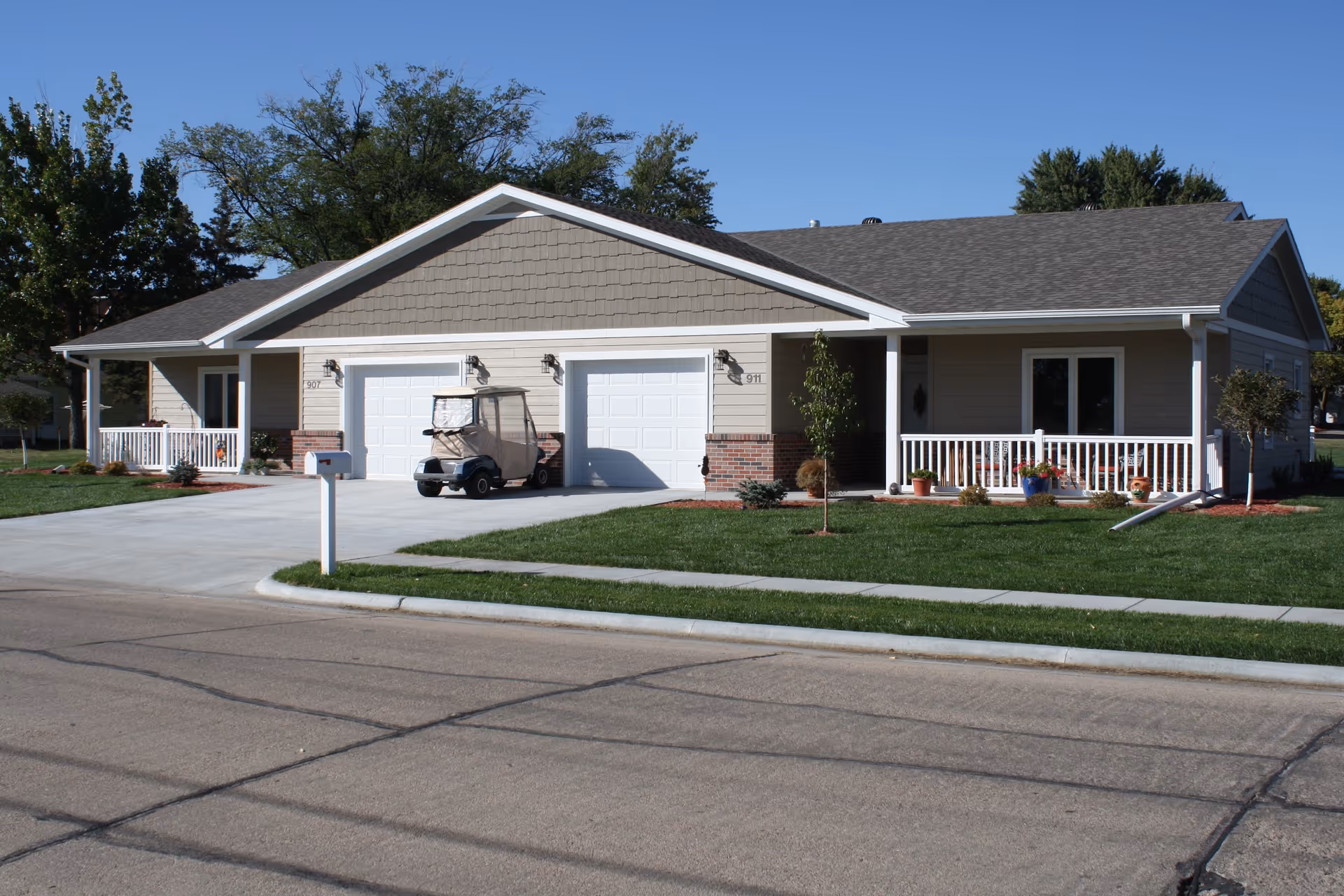 Exterior view of a single-story residential building with two white garage doors, a driveway, a golf cart parked in front of one garage, a small porch with white railings, green lawn, and trees in the background under a clear blue sky.