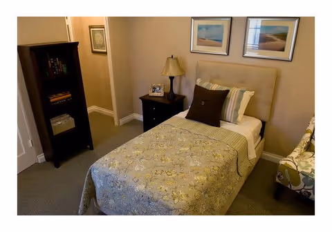 A cozy bedroom with a single bed covered in a floral-patterned quilt and multiple pillows. Next to the bed is a dark wooden nightstand with a lamp and a framed photo. On the wall above the bed are two framed pictures. To the left is a tall dark wooden bookshelf with books and decorative items. The room has beige walls and carpeted floor.