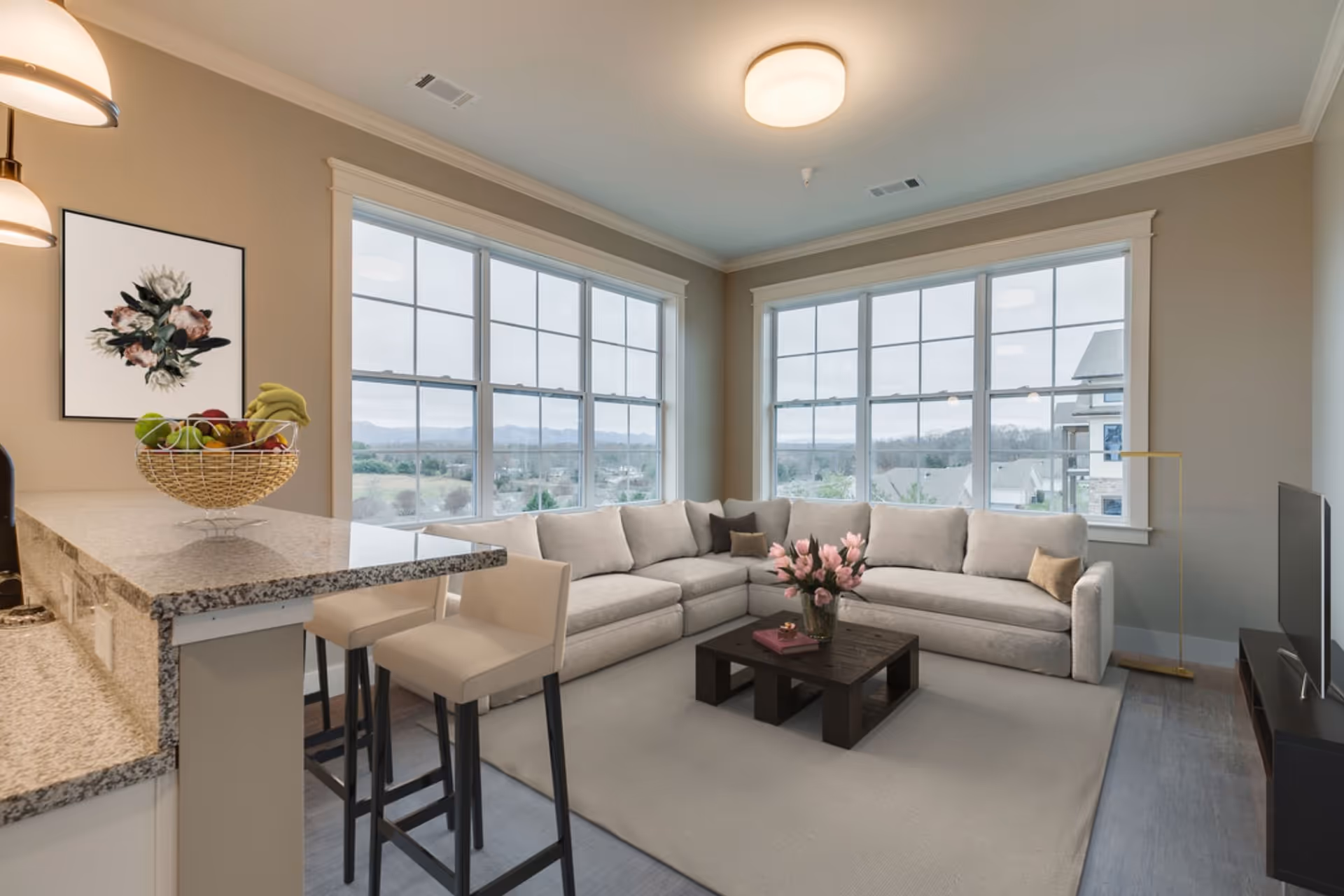 A bright living room with large windows showing an outdoor view. The room features a light gray L-shaped sectional sofa with several throw pillows, a dark wooden coffee table with a vase of pink flowers and books, a flat-screen TV on a black TV stand, and a kitchen counter with two beige bar stools and a fruit basket. The walls are painted beige, and the ceiling has a round light fixture.
