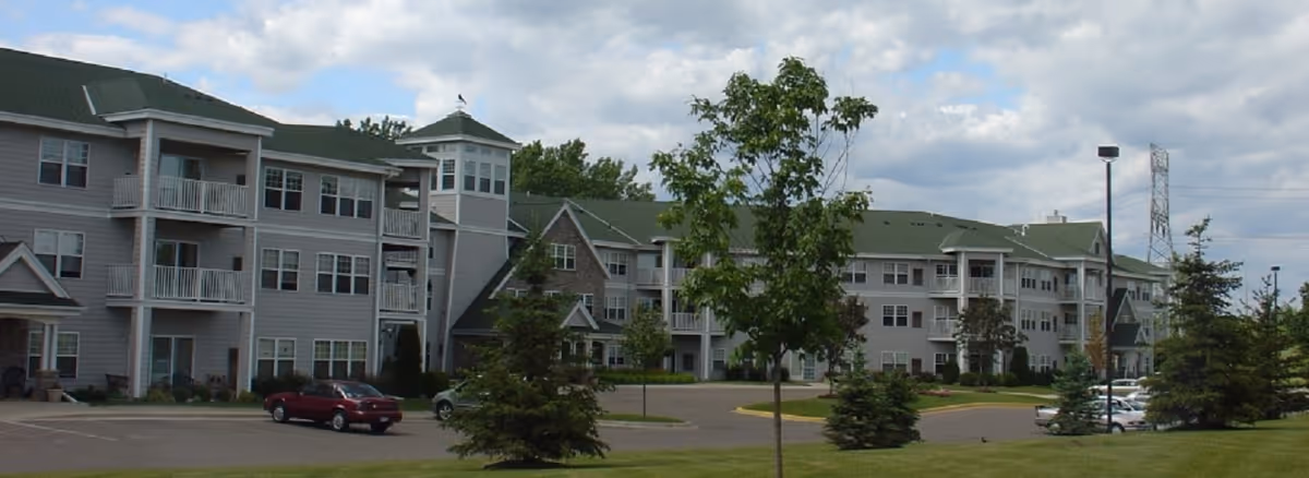 Exterior view of a multi-story senior living facility building with gray siding and green roofs, surrounded by trees, a parking lot with several cars, and a partly cloudy sky.