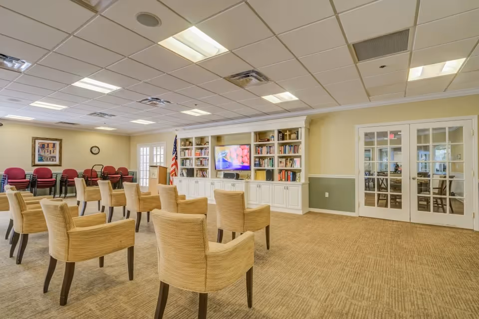 A spacious community room with beige carpet and walls painted in light yellow and green. The room features several beige armchairs arranged in rows facing a built-in white bookshelf with a flat-screen TV in the center. There is an American flag next to the bookshelf, a clock on the wall, and a stack of red chairs against the far wall. Double glass doors lead to another room with tables and chairs.