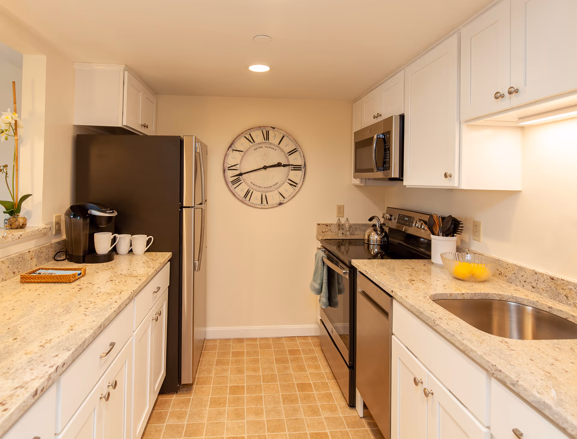 A modern kitchen with white cabinets, granite countertops, and stainless steel appliances including a refrigerator, microwave, stove, and dishwasher. There is a large round wall clock with Roman numerals on the far wall. On the left countertop, there is a coffee maker with three white mugs, and on the right countertop, there is a bowl with lemons and a utensil holder. The floor has a beige tile pattern.