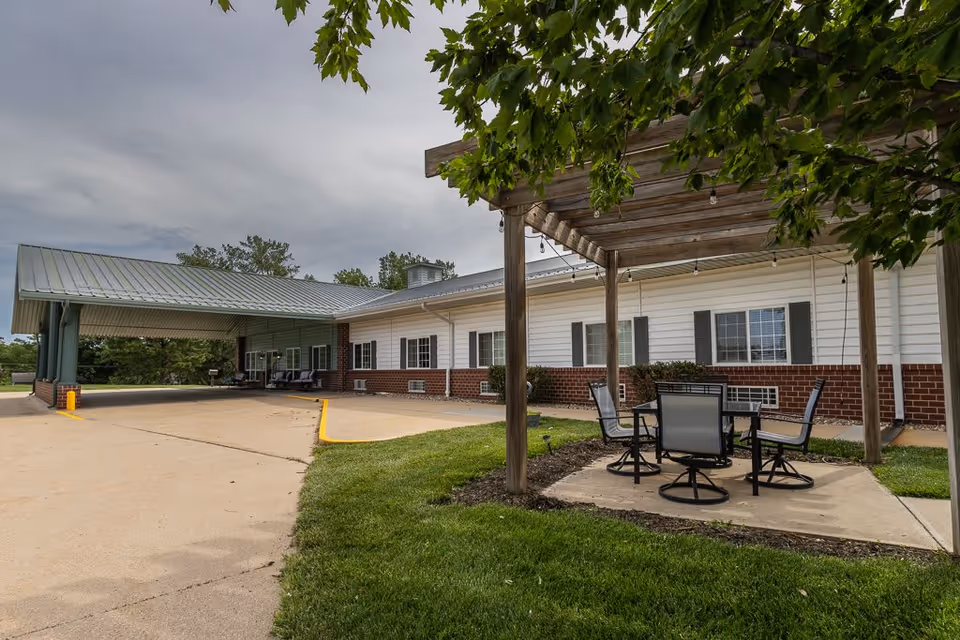 Outdoor seating area with a wooden pergola and four chairs around a table on a concrete patio, adjacent to a building with white siding and red brick accents. The building has several windows with black shutters and a covered driveway entrance. Green grass and trees surround the area under a cloudy sky.