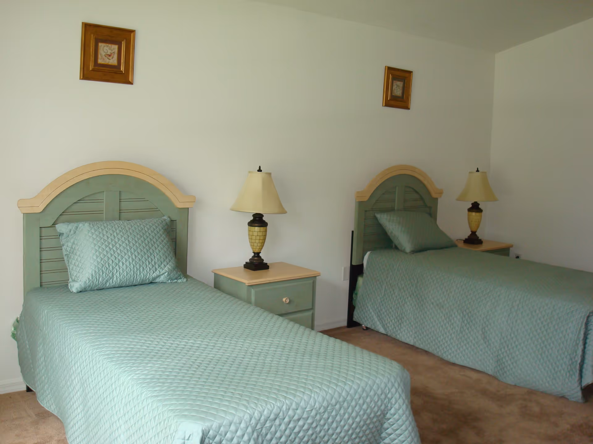 A bedroom with two twin beds, each with a light green quilted bedspread and matching pillow. Between the beds is a small nightstand with a lamp. The walls are white with two small framed pictures hanging above each bed.