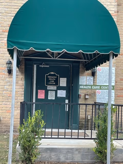 Entrance to Treemont Health Care Center with a green door under a green awning. The door has multiple signs including one that reads 'Treemont Health Care Center' and another indicating 'Ambulance'. There is a black metal railing in front of the entrance and some small bushes on either side. The building is made of light-colored brick.
